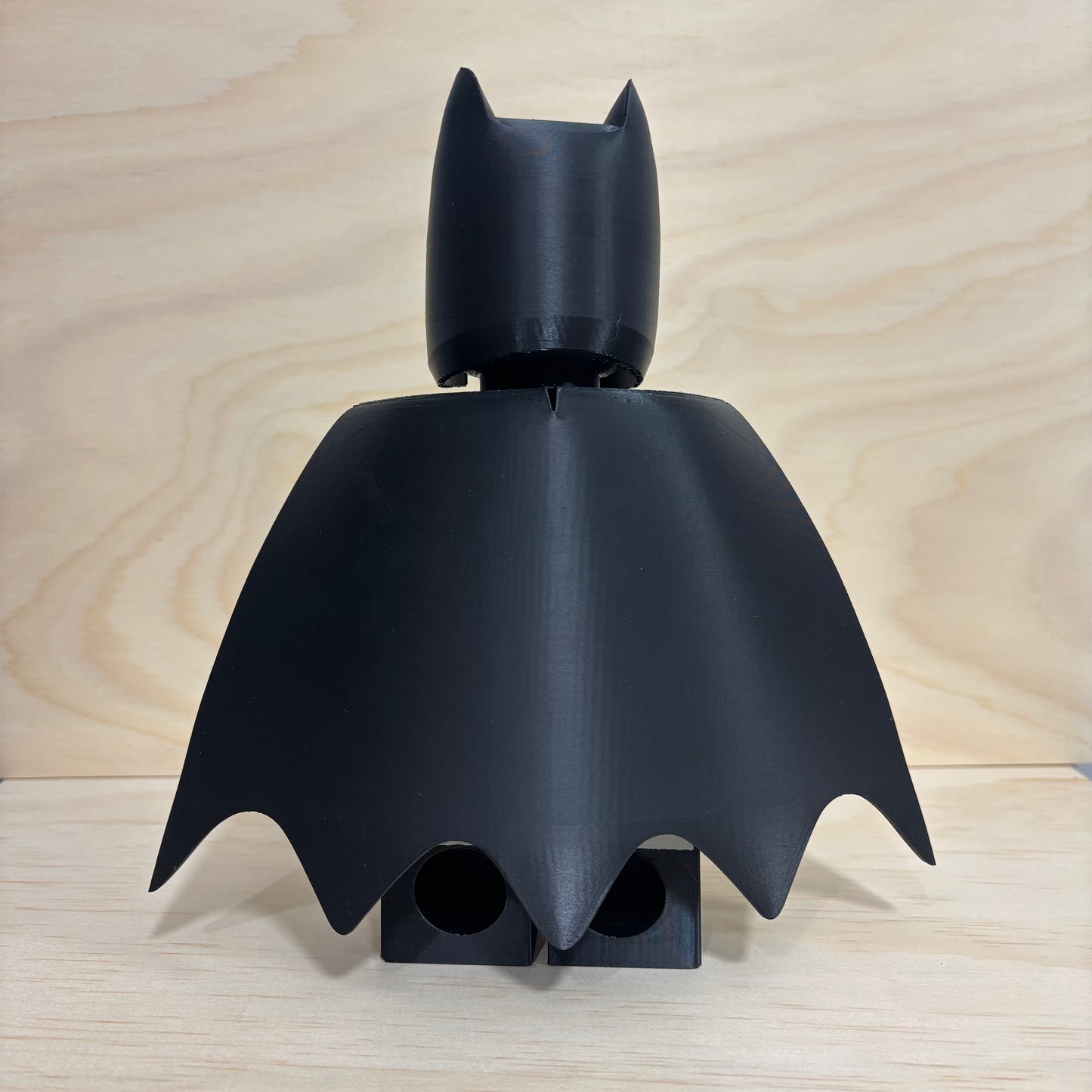 A matte black Batman-themed 3D-printed accessory, likely a toy minifigure head and cape assembly, displayed on a light wood surface against a matching wood grain background.