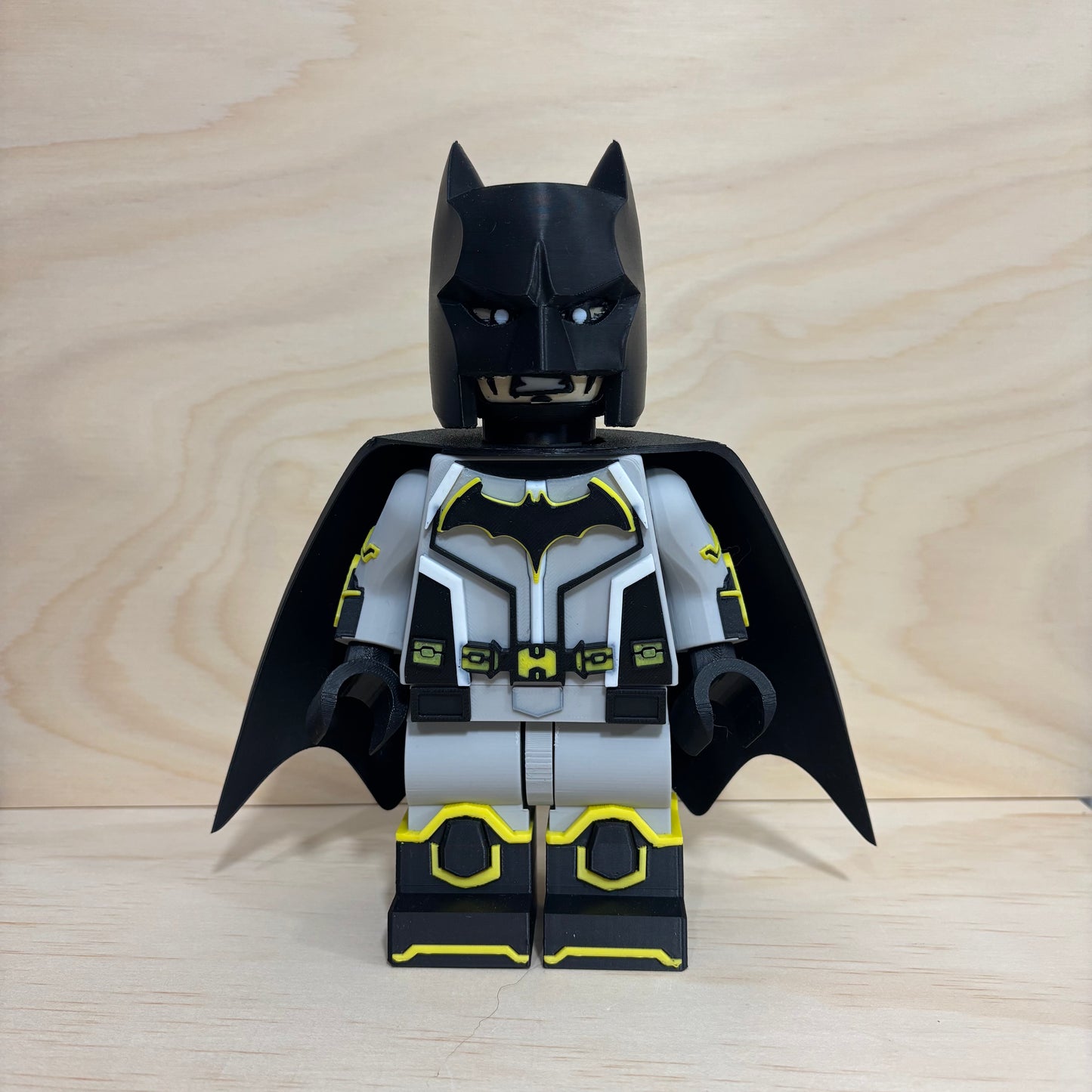 A custom-built LEGO Batman minifigure with a black and gray suit, yellow accents, and a black cowl, standing against a light wood grain background.
