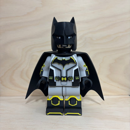 A custom-built LEGO Batman minifigure with a black and gray suit, yellow accents, and a black cowl, standing against a light wood grain background.